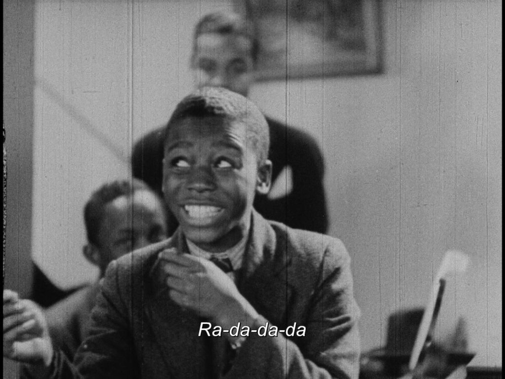 A Black boy in a suit mimics playing a trombone, with a big entertaining smile. Subtitles: Ra-da-da-da