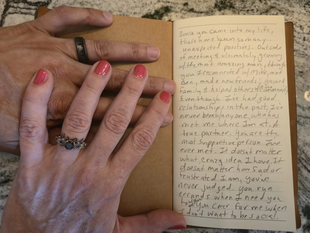 His and her hands, showing their wedding rings. They hold open the first page of her vows.