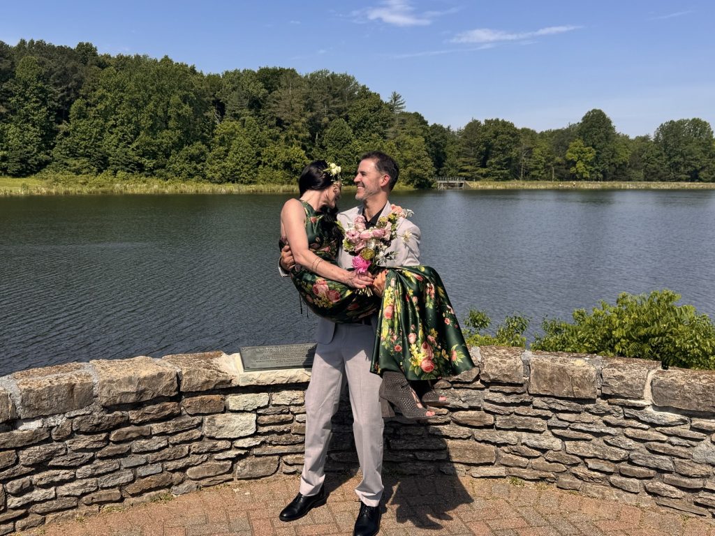 A newly-wed man in a gray suit carries his bride in his arms. She is wearing a green flowered dress. They stand in front of a small rock wall overlooking a lake on a sunny day.