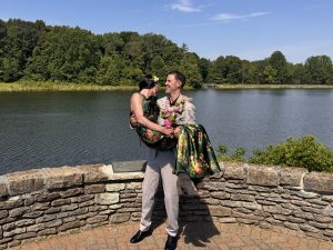 The groom holds his new wife in his arms in front of a lake