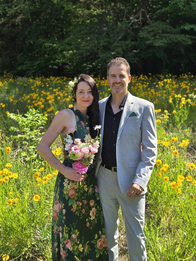 A newlywed couple stands in a field of yellow flowers. She is dressed in green with flowers in her hand and hair. He is in a gray suit with a green pocket square and black shirt.
